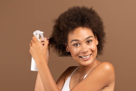 Téléchargez les photos : Smiling young african american curly female with perfect skin in white top applying spray on her hair, isolated on brown background, studio. Beauty care, cosmetics and styling, daily routines, haircut - en image libre de droit