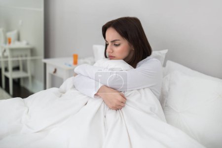 Foto de Mala joven morena bonita mujer en pijama blanco sentado en la cama bajo cubierta en casa, abrazando sus rodillas, mirando el espacio de copia, que sufre de resfriado, gripe, infección por coronavirus - Imagen libre de derechos
