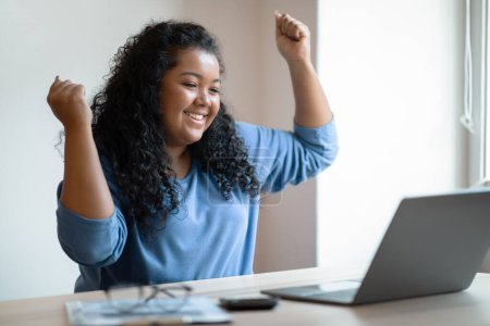 Foto de Emotional happy young chubby hispanic lady entrepreneur sitting at table, working from home, using laptop, looking at computer screen, gesturing and screaming, having good news, copy space - Imagen libre de derechos