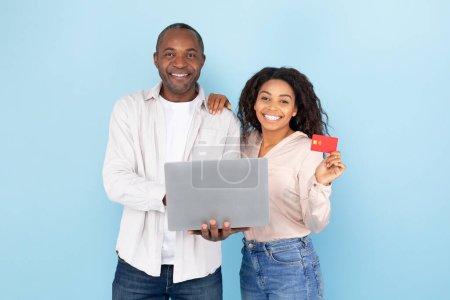 Photo for Happy black middle aged man and young woman showing credit card and laptop, standing over blue background, studio shot. Sale for shopaholics and order purchases, pay money and online shopping - Royalty Free Image