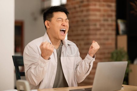Foto de Alegre hombre asiático de mediana edad utilizando el ordenador portátil y el gesto SÍ, sintiéndose feliz por ganar en línea o promoción de negocios en casa. Masculino lotería ganadora o apuesta de casino, juegos de azar en PC - Imagen libre de derechos