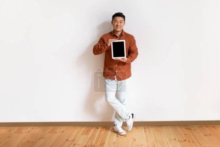 Photo for Happy middle aged asian man showing digital tablet with blank screen, standing against white wall, mockup for website or app, full length. Male demonstrating touch pad display template - Royalty Free Image