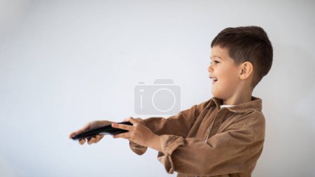 Foto de Smiling caucasian little child with remote control switches channels on TV set, chooses cartoon on white wall background, copy space. Boy enjoys free time, has fun, entertainment and watching videos - Imagen libre de derechos