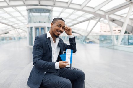 Téléchargez les photos : Happy Black Businessman Posing With Passport And Tickets In Hand At Airport, Handsome African American Male Sitting On Bench At Terminal Hall And Looking At Camera, Waiting For Flight, Copy Space - en image libre de droit
