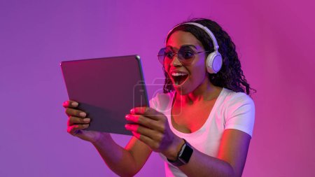 Téléchargez les photos : Portrait of Amazed Black Female In Headphones and Stylish Eyeglasses Using Digital Tablet, femme afro-américaine surprise écoutant de la musique ou jouant à des jeux vidéo tout en se tenant debout dans la lumière du néon - en image libre de droit