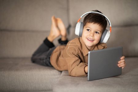 Foto de Smiling caucasian little kid in headphones enjoys free time, watching video, playing game on tablet, lying on sofa, looking at empty space. Study at home remotely, childhood and app for learning - Imagen libre de derechos