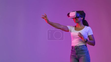 Photo for Virtual Experience. Smiling Black Lady Touching Air While Playing Video Game In VR Headset, Excited Young African American Woman Standing In Neon Light Over Purple Studio Background, Panorama - Royalty Free Image