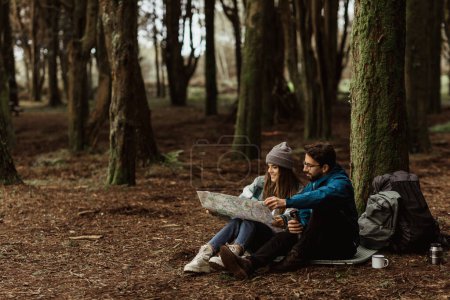 Téléchargez les photos : Smiling young european man and woman tourists in jackets with backpack resting in autumn forest, enjoy cold season outdoors, looking at map with hot drink from thermos. Route for travel and nature - en image libre de droit