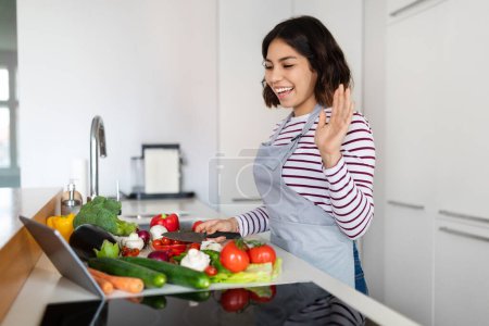 Téléchargez les photos : Cool friendly cheerful pretty brunette young woman food blogger wearing apron streaming while cooking healthy tasty meal at cozy modern kitchen, waving at digital pad screen, copy space - en image libre de droit