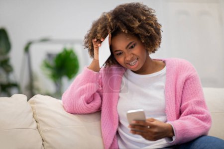 Téléchargez les photos : Confused millennial curly african american woman sitting on couch in living room, holding correspondence from bank, looking at phone screen, female customer banking online, have financial problems - en image libre de droit