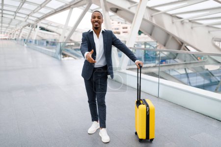 Foto de Smiling young black businessman extending hand for handshake at camera while standing in modern airport terminal, african american male entrepreneur in suit greeting somebody, copy space - Imagen libre de derechos
