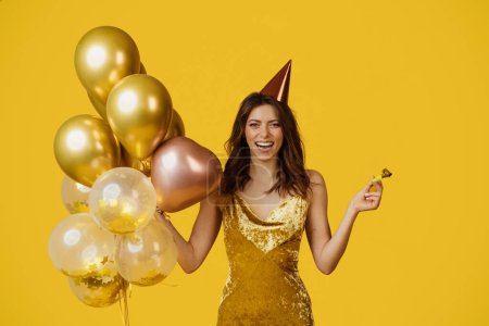 Photo for Overjoyed lady with festive balloons, birthday cap and party blower celebrating special occasion over yellow studio background. Happy woman having fun on holiday - Royalty Free Image