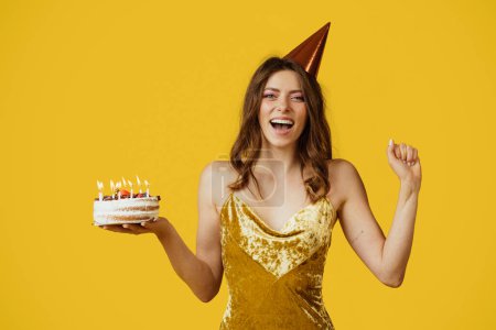 Photo for Overjoyed woman in dress and birthday hat holding cake, posing isolated on yellow studio background. Emotions from celebrating holiday and make wish for birthday - Royalty Free Image