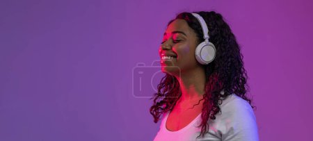 Téléchargez les photos : Smiling Relaxed Black Lady Listening Music In Wireless Headphones, Happy Young African American Woman Standing With Closed Eyes In Neon Light Over Purple Background, Enjoying Favorite Song, Panorama - en image libre de droit