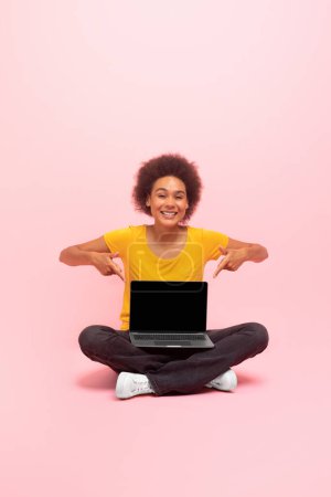 Téléchargez les photos : Cheerful young african american curly woman sit on floor, point fingers at laptop with blank screen recommend ad, offer isolated on pink background. Advice for work and study remotely, device and blog - en image libre de droit
