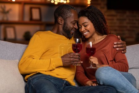 Téléchargez les photos : Loving handsome middle aged black husband hugging pretty millennial wife, cheerful smiling couple cuddling on sofa, drink wine in the evening after job, home interior. Love, relationhships, marriage - en image libre de droit
