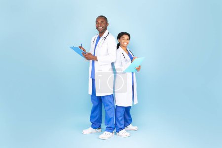 Foto de African american man and woman doctors in white coats with stethoscopes writing at tablet and smiling, blue background, full length. Medical service, examination with therapist, prescription - Imagen libre de derechos