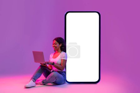 Téléchargez les photos : Smiling black lady with laptop leaning at big blank smartphone with white screen while sitting in neon light over purple studio background, happy female browsing new website, collage, mockup - en image libre de droit