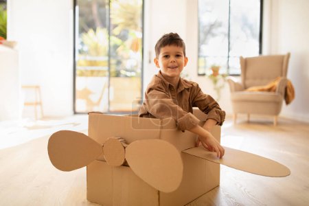 Téléchargez les photos : Cheerful cute caucasian little child sit in cardboard box airplane, has fun alone, enjoy travel in living room interior, sun flare. Entertainment and play game at home, fantasy, art and childhood - en image libre de droit