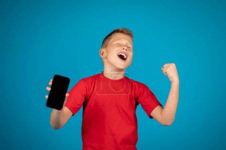 Photo for Overjoyed boy holding blank smartphone and celebrating success while standing isolated over blue background, excited male kid cheering online game win, shaking fist and screaming with joy, mockup - Royalty Free Image