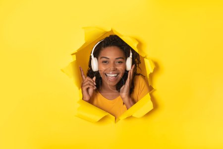 Photo for Happy black lady enjoying music in headphones posing and smiling at camera through hole in torn yellow paper background, studio shot. Collage - Royalty Free Image
