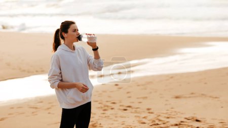 Foto de Deporte e hidratación. Mujer en forma joven bebiendo agua mineral de la botella, descansando después de entrenar en la playa junto al mar, panorama con espacio para copiar - Imagen libre de derechos