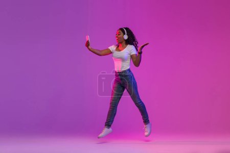 Foto de Mujer negra alegre en luz de neón saltando con el teléfono inteligente en las manos, Mujer afroamericana alegre haciendo videollamada o navegando por la aplicación en el teléfono móvil sobre fondo de gradiente púrpura, espacio de copia - Imagen libre de derechos