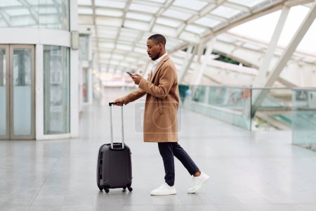 Téléchargez les photos : Online Check-In. Young Black Man Walking With Suitcase In Airport And Using Smartphone, African American Male Browsing Internet Or Messaging On Cellphone While Going To Flight Gate, Copy Space - en image libre de droit