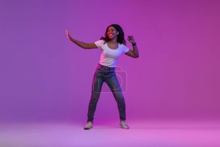 Téléchargez les photos : Jeune femme noire insouciante dansant dans la lumière au néon dans le studio, pleine longueur prise de vue d'une belle femme afro-américaine heureuse faisant des mouvements de danse et s'amusant sur fond violet dégradé, Copier l'espace - en image libre de droit