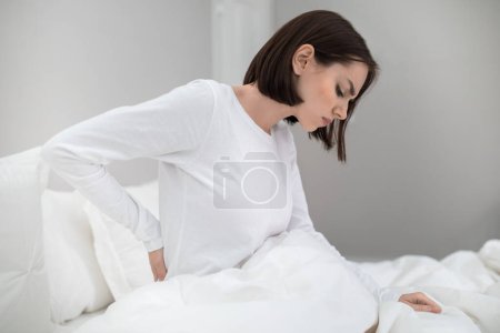 Téléchargez les photos : Jeune femme malheureuse souffrant de maux de dos après avoir dormi dans la chambre, se réveillant le matin, assise sur le lit et touchant le bas du dos, portant un pyjama blanc, espace de copie, vue latérale - en image libre de droit