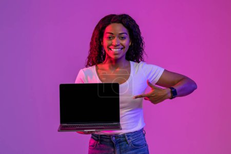 Foto de Anuncio en línea. Mujer afroamericana feliz apuntando a la pantalla de computadora portátil en blanco, sonriendo hembra negra recomendando un nuevo sitio web mientras está de pie en la iluminación de neón sobre fondo púrpura, burla - Imagen libre de derechos