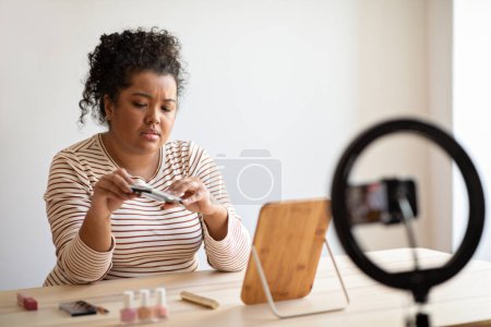 Photo for Unhappy young hispanic woman plus size beauty blogger recording video for followers, sitting at desk full of cosmetics, holding eyeshadow palette, streaming from home, using blogger set, copy space - Royalty Free Image