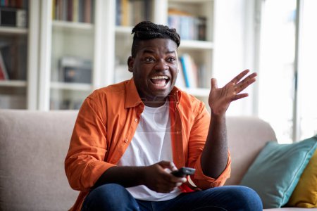 Foto de Exciting happy overweight african american millennial guy with dreadlocks hairstyle sitting on couch at home, switching tv, watching nice film or show, free space. Domestic entertainment concept - Imagen libre de derechos