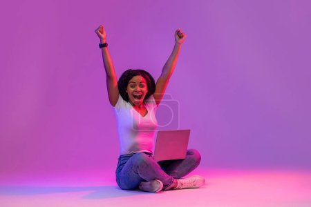 Photo for Euphoric Black Female With Laptop Celebrating Success In Neon Light, Excited Young African American Woman With Computer Sitting On Floor Over Purple Studio Background And Raising Hands, Copy Space - Royalty Free Image