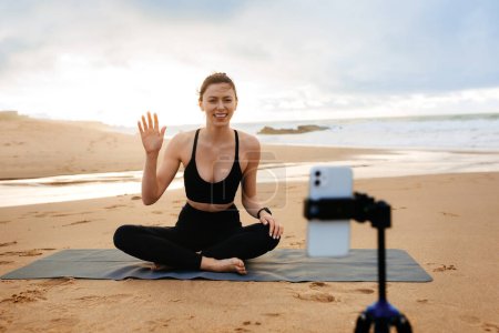Foto de Online training. Sporty woman yoga coach recording video online training, sitting on fitness mat on the beach by seaside. Sport, active life, blogging - Imagen libre de derechos