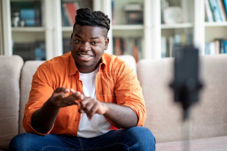 Photo for Cheerful handsome overweight young black guy in casual outfit with braids famous blogger or influencer sitting on couch, looking at phone set on tripod, gesturing and smiling, streaming from home - Royalty Free Image