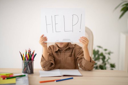 Photo for Bored upset tired caucasian little child sits at table shows paper with inscription help, suffer from overwork in room interior. Problems at study, education at school and home, childhood and lesson - Royalty Free Image