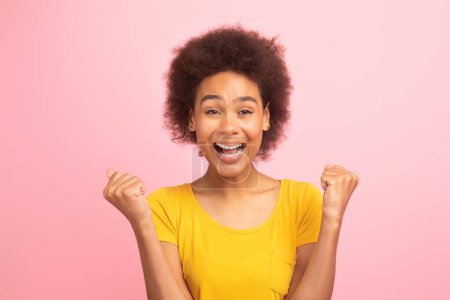 Téléchargez les photos : Joyeux excité jeune afro-américaine bouclée dame en t-shirt jaune à bouche ouverte faire geste de victoire et de succès avec les mains, regarder la caméra isolée sur fond rose, studio. Bonne chance émotion - en image libre de droit