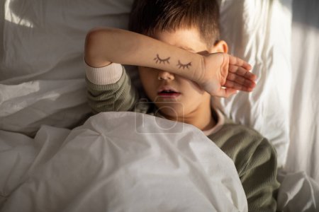 Foto de Tired peaceful caucasian little child sleeps on comfortable bed covers his face with hand with drawn closed eyes in bedroom, close up, top view. Good sleep, health care, rest and relaxation at night - Imagen libre de derechos