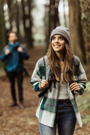 Téléchargez les photos : Smiling inspired young european guy and lady tourists in jackets, hat with backpack walk in autumn forest, enjoy cold season outdoor, blurred, vertical. Travel, adventure lifestyle and relationships - en image libre de droit