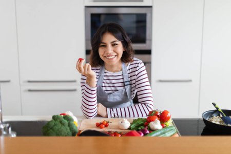 Foto de Mujer hispana bastante joven y feliz usando delantal cocinando en casa, sonriente dama mostrando verduras orgánicas frescas de tomate rojo cereza, preparando comida saludable, interior de la cocina, espacio para copiar - Imagen libre de derechos