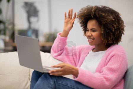 Photo for Cheerful smiling attractive bushy young black woman waving at laptop screen, sitting on couch in living room, have online meeting with boyfriend or family, home interior, copy space - Royalty Free Image