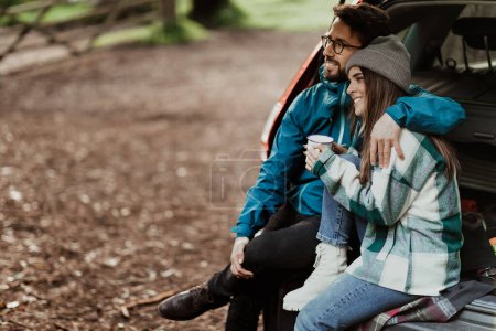 Foto de Sonriendo joven pareja europea turistas en chaquetas relajarse en el bosque de otoño, disfrutar de una taza de bebida caliente y sentado en el maletero del coche en el tiempo libre. Viajar juntos, estilo de vida activo, aventura y relación - Imagen libre de derechos