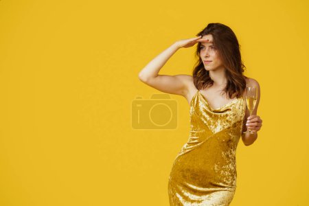 Téléchargez les photos : Jeune femme en robe de soirée élégante tenant un verre de champagne et regardant de côté, tenant la main au front isolé sur fond de studio jaune, espace de copie - en image libre de droit