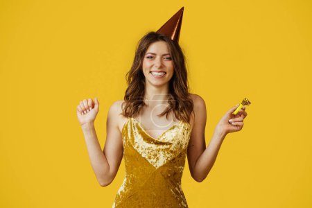 Téléchargez les photos : Excited lady in dress and birthday cap holding party horn, celebrating her special day on yellow studio background. Gorgeous young lady having holiday fun - en image libre de droit