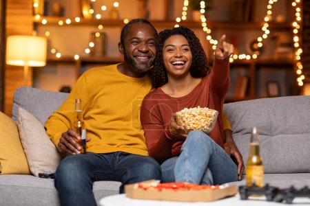 Téléchargez les photos : Amoureux africains américains heureux émotionnels profitant d'une belle soirée ensemble à la maison, homme et femme noirs joyeux regarder un film, manger de la pizza, pop-corn, boire de la bière, dame excitée pointant vers l'espace de copie - en image libre de droit