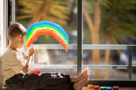 Photo for Busy caucasian little child sits on floor with paints, draws rainbow on panoramic window in elementary school room interior. Fun alone, entertainment and play at home, art, fantasy and childhood - Royalty Free Image