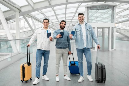 Téléchargez les photos : Vacation With Friends. Three Travelers Men Holding Boarding Pass Tickets And Passports Traveling Abroad Together Standing With Suitcases Smiling To Camera In Modern Airport Indoors - en image libre de droit