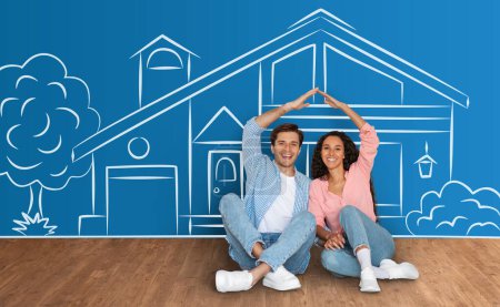 Photo for Cheerful millennial couple sitting on floor, holding hands above their heads, showing roof, posing over nice house sketch on blue background, collage. Happy family moving to new apartment - Royalty Free Image