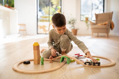 Foto de Smiling european small kid in pajamas plays with toys, train, cars and wooden road, sits on floor in bright living room interior. Fun alone, childhood and entertainment, fantasy at home, kindergarten - Imagen libre de derechos